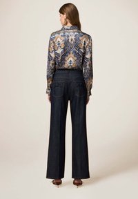Blusa de manga larga en un patrón de paisley azul y dorado, combinada con pantalones de mezclilla anchos oscuros que cuentan con dos bolsillos traseros y acentos de lazo.