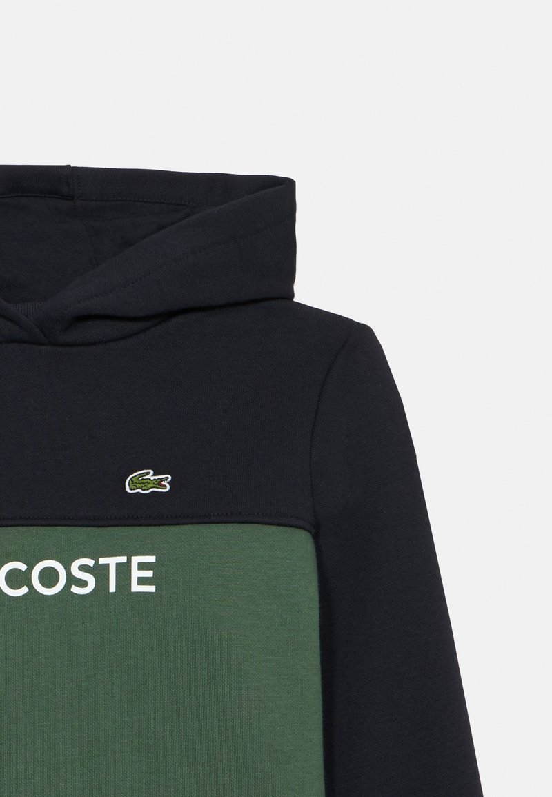 Sweat à capuche en vert et bleu marine, comportant un grand logo "LACOSTE" blanc et un petit emblème crocodile brodé. Texture de tissu lisse.