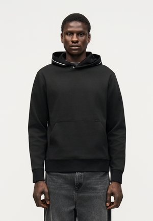 Mężczyzna ubrany w zwykłą czarną bluzę z kapturem i kieszenią z przodu oraz wyblakłe czarne dżinsy, stojący na białym tle.