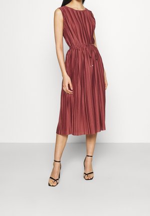 Femme portant une robe mi-longue sans manches plissée de couleur rouille avec une ceinture à la taille, associée à des sandales noires à talons hauts et à brides.