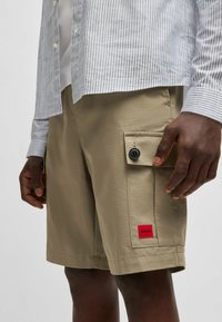 Pantaloni cargo beige realizzati in tessuto testurizzato, dotati di una tasca laterale con chiusura a bottone e di un'etichetta rossa con il marchio sull'orlo.