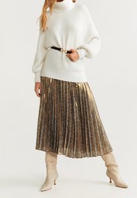 Chunky witte gebreide coltrui gecombineerd met een geplooide, glanzende gouden en zwarte rok,accentueert met een zwarte riem en beige knielange laarzen.