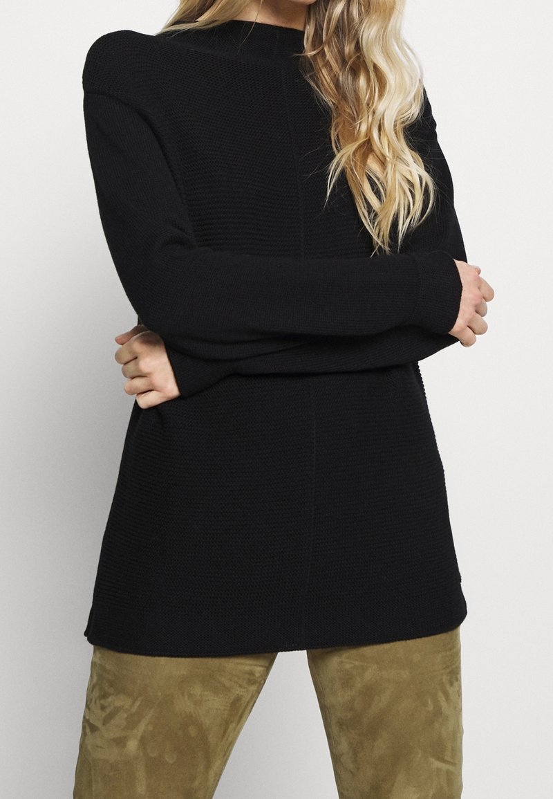 Pull en maille noir à manches longues, tissu texturé et coupe décontractée, associé à un pantalon en daim vert clair.