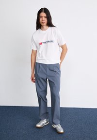 Camiseta blanca con logo rojo y azul, combinada con pantalones grises y zapatillas deportivas plateadas. El atuendo presenta un corte holgado y un diseño casual.
