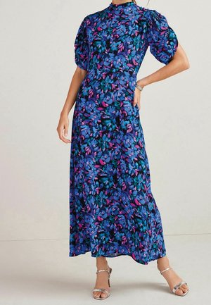 Kobieta w długiej kwiecistej sukience z niebieskimi, fioletowymi i różowymi kwiatami, z bufiastymi krótkimi rękawami i srebrnymi sandałami na wysokim obcasie.