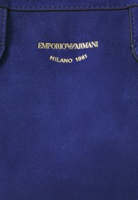 Modra nubuk tkanina z zlatim vtisnjenim napisom "EMPORIO ARMANI MILANO 1981" ter logotipom v središču.