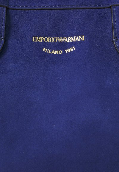 Mėlynas zomšinis audinys su aukso įspaustu tekstu "EMPORIO ARMANI MILANO 1981" ir logotipu centre.