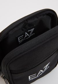 Czarna saszetka na zamek błyskawiczny z teksturowanym wykończeniem, z naszywką logo w środku i gładkim podszewką. Kompaktowy i prostokątny kształt.