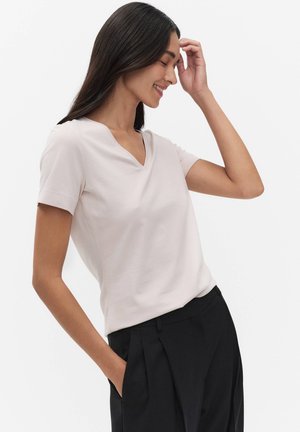 Vrouw met lang donker haar, gekleed in een lichtroze V-hals T-shirt met korte mouwen en zwarte geplooide broek, glimlachend met één hand die haar hoofd aanraakt.