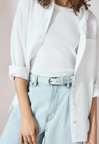 Chemise blanche à boutons, débardeur blanc côtelé, jean taille haute bleu clair et ceinture en cuir bleu pastel avec boucle en argent.