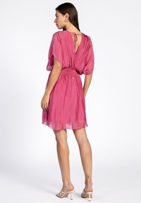 Korte roze jurk met een vloeiende stijl, geplooide achterkant, elastische taille en striksluiting bij de hals. Transparante textuur en boven de knie lengte.