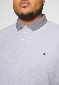 Šedé polo tričko s texturovanou tkaninou. Má černobílý texturovaný límeček, dva knoflíky a malé vyšívané logo na hrudi.