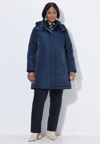 Parka aislante azul marino con capucha, que cuenta con un cierre frontal, dos bolsillos laterales y un corte más largo, llevada sobre pantalones azul marino y tacones beige.