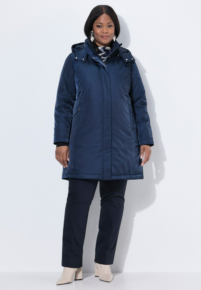 Parka aislante azul marino con capucha, que cuenta con un cierre frontal, dos bolsillos laterales y un corte más largo, llevada sobre pantalones azul marino y tacones beige.