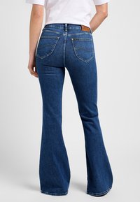 Uitlopende blauwe denim jeans met een hoge taille, stiksel details en twee achterzakken. De stof heeft een gestructureerde afwerking en minimale slijtage.