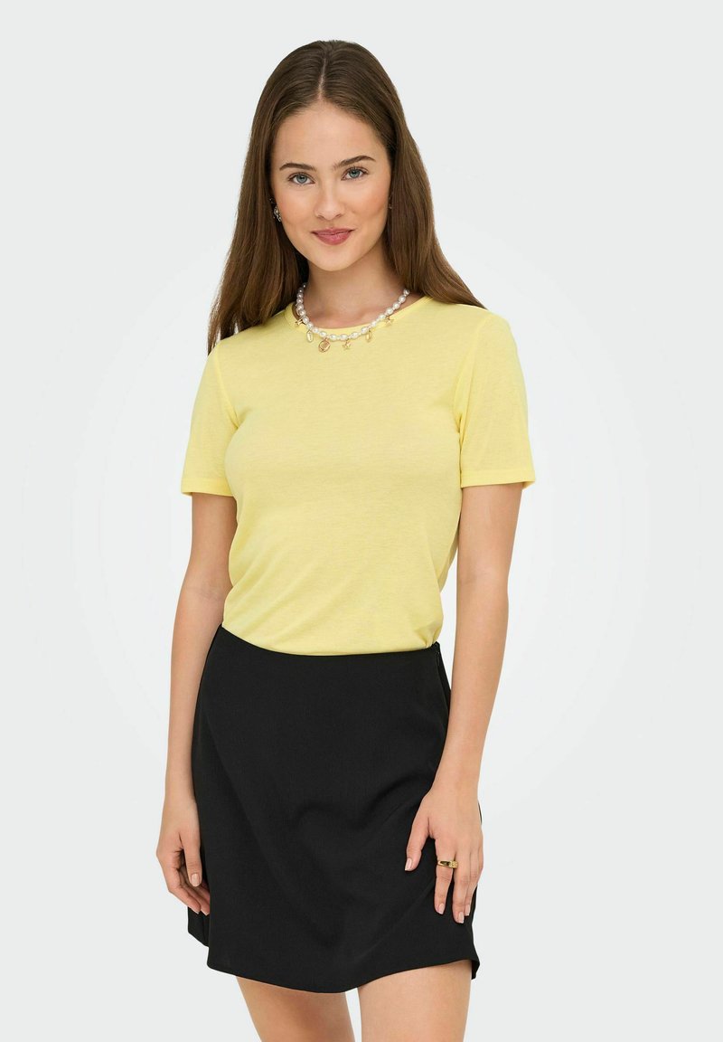 Młoda kobieta z długimi brązowymi włosami, ubrana w żółtą koszulę z krótkim rękawem, czarną spódnicę, naszyjnik z pereł i złoty pierścionek, stojąca na białym tle.