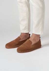 Chaussures à enfiler en suède brun avec une semelle en jute tissée, dotées d'un dessus lisse et de côtés élastiques pour un enfilage facile.