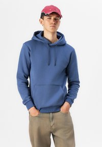 Sudadera azul con un bolsillo frontal, cordón ajustable y puños acanalados, combinada con pantalones marrón claro y una gorra roja desgastada.