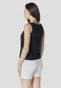 Femme aux longs cheveux noirs portant un haut noir sans manches et un short en denim blanc, debout de profil devant un fond uni.