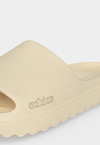 Sandale slide beige Adidas avec une semelle épaisse et un logo embossé sur le côté, conçue pour un usage décontracté et confortable.