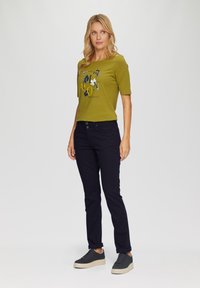 Camiseta verde de manga corta con un gráfico impreso en el frente, combinada con pantalones oscuros azul marino y zapatillas negras con una suela texturizada.