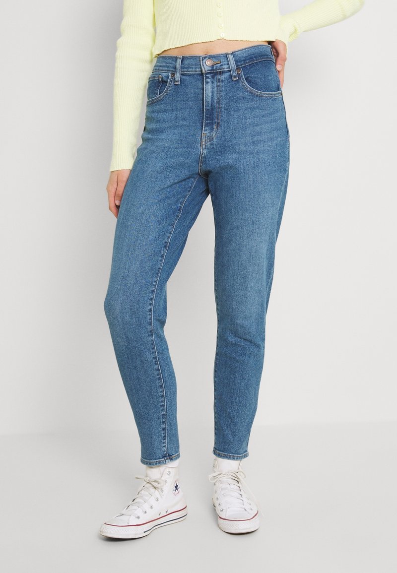 Personne portant un jean bleu taille haute, un pull court jaune pâle, des baskets blanches, debout devant un fond clair uni.