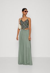 Top crop floral com alças finas, combinado com uma saia maxi longa e plissada de cor verde-clara. A modelo segura uma pequena mala branca e usa óculos de sol rosa.