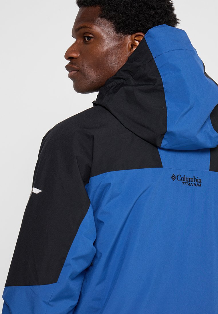 Mann, der eine blaue und schwarze Columbia Titanium Jacke mit hochgeklappter Kapuze trägt, blickt nach links vor einem einfachen hellen Hintergrund.