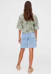 Haut vert à manches courtes effet tie-dye associé à un short en denim bleu clair avec ourlets effilochés et poches arrière, porté avec des baskets beiges.