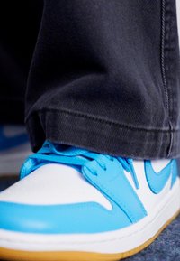 Chaussure de sport bleue et blanche avec une semelle en caoutchouc, design à lacets et accents cousus. Un pantalon en denim noir couvre une partie de la chaussure.