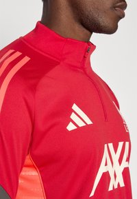Rød atletisk zip-up jakke med et Adidas-logo og hvid branding. Glat tekstur med kontrasterende orange accenter på skuldrene.