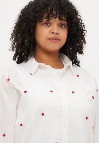 Λευκό πουκάμισο με κουμπιά και μικρές κόκκινες κεντητές καρδιές. Διαθέτει κλασικό γιακά και μακριά μανίκια, φτιαγμένο από ελαφρύ ύφασμα.