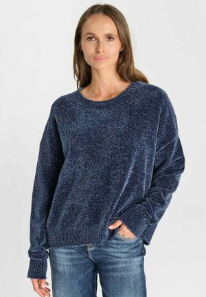 Vrouw die een losse donkerblauwe gebreide trui en blauwe jeans draagt, staand met één hand in haar zak tegen een egale lichte achtergrond.