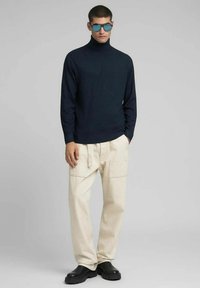 Maglione a collo alto blu navy, pantaloni cargo beige con tasche laterali e scarpe nere. Il modello indossa occhiali da sole specchiati, in piedi su sfondo neutro.