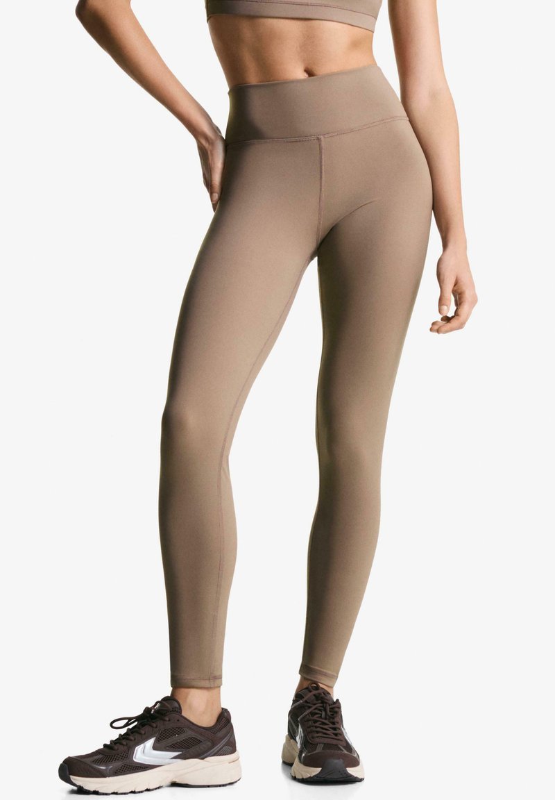 Højtsiddende, brune sømløse leggings lavet af strækstof med en glat tekstur, parret med mørke atletik sko.