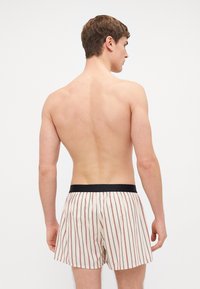Boxer shorts rayés en crème avec des lignes rouges. Taille élastique noire. Tissu léger avec une texture lisse. Vue de dos du porteur.