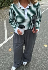 Green and white striped long-sleeve polo shirt, paired with loose gray trousers and white sneakers. Holding a coffee cup and a tablet.