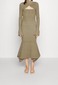 Femme portant une robe midi olive en tricot à manches longues avec des poignet évasés, une découpe au niveau de la poitrine et un ourlet asymétrique, accompagnée de sandales blanches.
