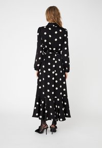 Abito nero con pois bianchi, maniche lunghe, vita cintrata, orlo fluido e fondo a balze, abbinato a tacchi neri e collant con motivo.