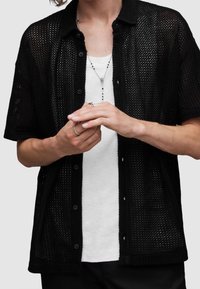 Camicia nera a maniche corte con bottoni, realizzata in un materiale a rete testurizzato, sovrapposta a una t-shirt bianca, con una vestibilità rilassata e bottoni in vista.
