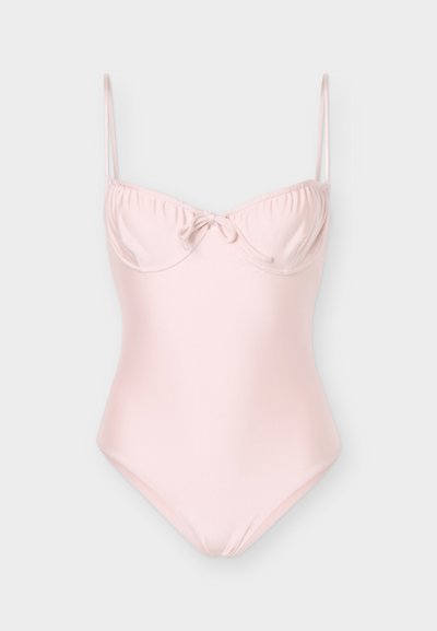 Jasnoróżowy jednoczęściowy strój kąpielowy z cienkimi, regulowanymi ramiączkami i małą, zawiązaną kokardką na środku marszczonej części biustu.