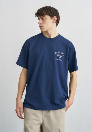 Joven con cabello corto y oscuro, vistiendo una camiseta azul marino de Carhartt WIP y pantalones cortos cargo beige, mirando hacia su izquierda.