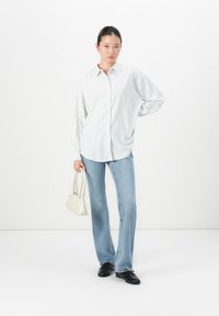 Kvinne står iført en løs hvit stripete skjorte, lyseblå jeans, svarte sko, og holder en liten hvit håndveske ved sin side.