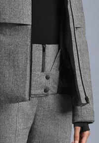 Detailný záber na osobu v šedom sako s vzorom rybej kosti a nohaviciach so zipsovými a cvokovými detailmi, a čiernou vnútornou vrstvou.