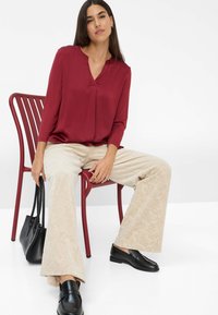 Una mujer se sienta en una silla roja, vistiendo una blusa burdeos, pantalones anchos beige con un patrón, mocasines negros y sosteniendo un bolso negro.
