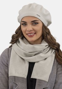 Beret tricotat de culoare gri deschis și eșarfă asortată cu o textură netedă, prezentând un model subtil cu dungi și un mic accent cu un logo metalic.