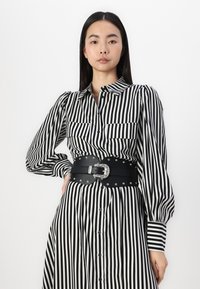 Μαύρο και άσπρο ριγέ φόρεμα πουκαμίσα με μακριά μανίκια puff, δύο τσέπες στο στήθος και μια φαρδιά μαύρη ζώνη με ασημένια μεταλλικά στοιχεία και καρφιά.