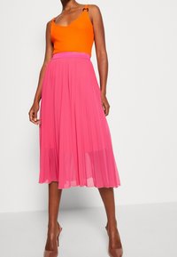 Top arancione senza maniche con spalline, abbinato a una gonna midi rosa plissettata. La gonna presenta una vita testurizzata e un tessuto fluido.