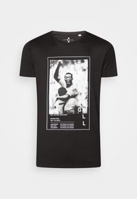 Camiseta de algodón negra con un gráfico en la parte frontal que presenta una imagen en blanco y negro de un hombre celebrando y un texto que detalla logros deportivos.