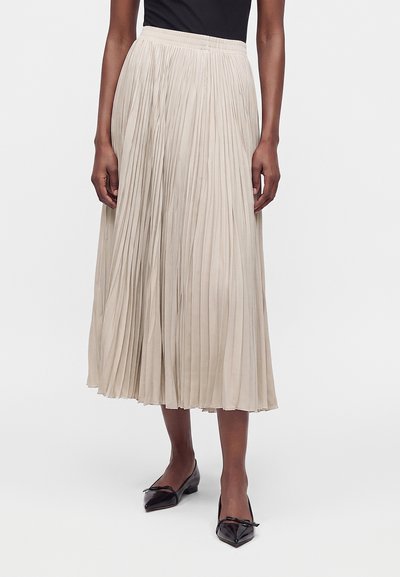 Femme portant une jupe midi plissée beige, un top noir et des ballerines noires à bout pointu avec petits nœuds, debout sur fond blanc.